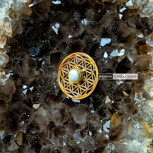 May include: Gold-toned metal charm with a flower of life design and a white opal stone in the center. The charm is on a dark gray background.