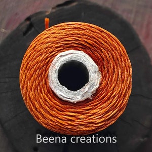 May include: A spool of orange thread wound around a white cardboard core. The thread is tightly wound and has a smooth, even texture.