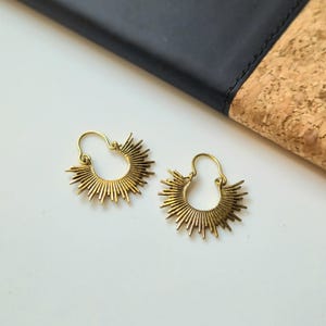 May include: A pair of gold-colored hoop earrings. Each earring features a sunburst design with radiating lines. The earrings are displayed on a white surface, with a black and cork-colored background.