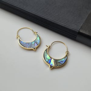 May include: A pair of gold-colored hoop earrings with a crescent moon design. The earrings feature iridescent abalone shell inlays, showcasing a mix of blues, greens, and purples. The earrings are displayed on a white surface.