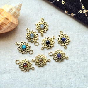 Puede incluir: Ocho dijes de metal dorado con un intrincado diseño calado de flores o mandalas. Cada dije tiene en el centro una pequeña piedra preciosa cabujón redonda y lisa en varios colores, incluyendo azul, morado, marrón y rojo oscuro.