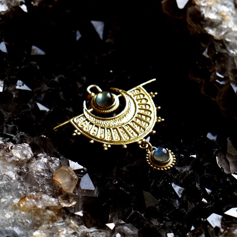 Puede incluir: Un colgante de oro con un dise&ntilde;o de media luna y dos piedras preciosas azul verdosas. El colgante cuelga de una cadena y se coloca sobre un fondo de cristales negros.