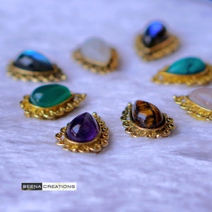 以下が含まれることがあります： さまざまな色の、緑、紫、茶色などの宝石がセットされた、しずく型のペンダントのコレクション。金色の装飾的な金属フレームに収められています。ペンダントは白いテクスチャのある表面に配置されています。「BEENA CREATIONS」のブランド名が表示されています。
