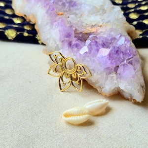 May include: A gold-coloured, openwork lotus flower-shaped pendant with an oval centre, next to two white seashells. The pendant is on a cream-coloured surface, with a large purple and white crystal formation in the background.