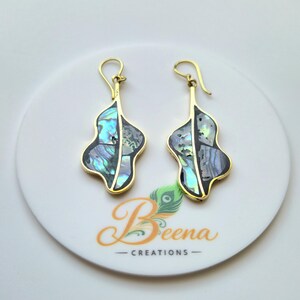 Pode incluir: Um par de brincos dourados com um design único em forma de folha. Cada brinco apresenta uma incrustação iridescente de concha de abalone em tons de azul, verde e cinza, emoldurada em ouro. O nome da marca "Beena Creations" é visível.