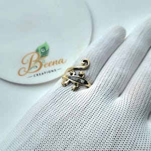 Puede incluir: Un anillo de metal dorado con un diseño de lagarto que presenta una incrustación oscura e iridiscente en el lomo. La cola del lagarto se curva hacia arriba y su cuerpo está detallado con pequeñas patas y pies sobre un fondo blanco.