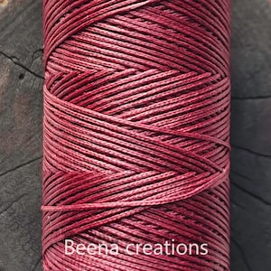 Könnte beinhalten: Eine Spule mit tiefrotem gewachstem Baumwollgarn. Das Garn ist fest gewickelt und hat eine glatte, polierte Oberfläche.