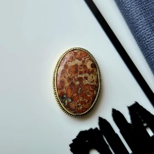 Puede incluir: Un broche ovalado que presenta una piedra pulida multicolor con un patrón moteado en tonos óxido, bronceado y crema. La piedra está engastada en un marco de metal dorado texturizado que tiene un detalle delicado de borde trenzado.