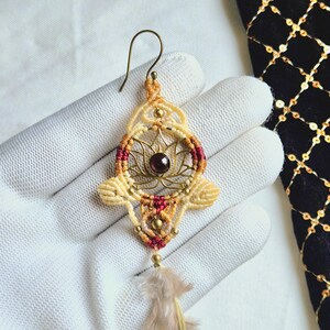 May include: A gold and cream macrame earring with a red and gold lotus design and a garnet gemstone. The earring has a feather charm hanging from the bottom.