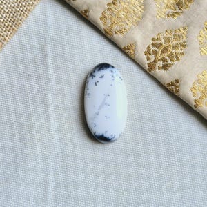 Puede incluir: Un cabujón ovalado blanco con inclusiones dendríticas negras. La piedra está colocada sobre un fondo de tela gris claro.