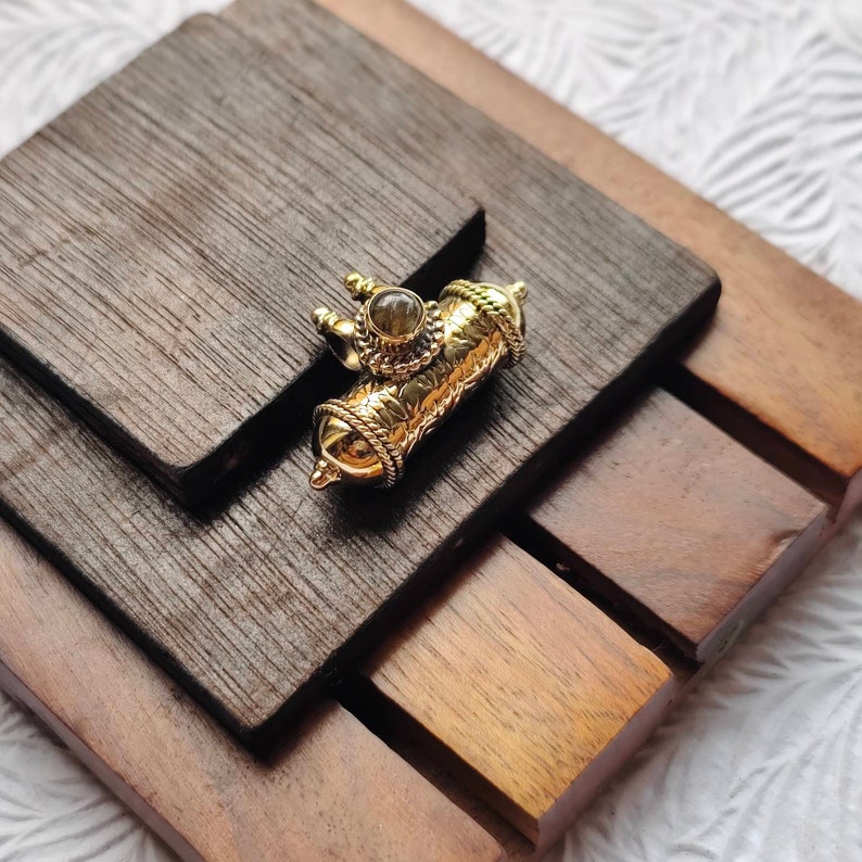 May include: A gold-toned pendant with intricate details and a clear gemstone in the center. The pendant is on a wooden surface.