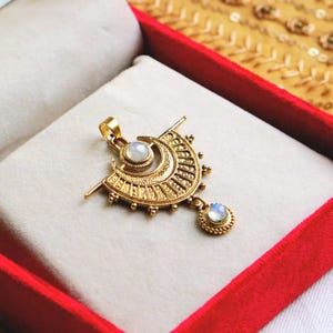 Puede incluir: Un colgante de oro con un diseño de media luna y dos gemas de piedra lunar iridiscente. El colgante está detallado con intrincados tallados y abalorios.