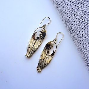 May include: A pair of gold-toned dangle earrings featuring a celestial motif. Each earring displays a sun and crescent moon at the top, with long, vertical rays extending downward, accented by small, cut-out star shapes throughout the design.
