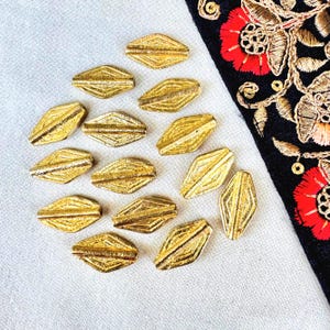 Puede incluir: Cuentas doradas en forma de diamante con superficies texturizadas. Las cuentas están dispuestas sobre una tela blanca, con una porción de tela negra con bordados florales rojos visibles. Adecuado para la fabricación de joyas.