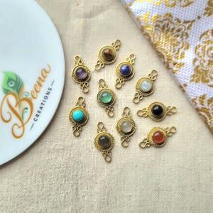 May include: Ten gold-toned metal jewellery connectors, each featuring a central round cabochon gemstone in various colours including turquoise, amethyst, lapis lazuli, and tiger's eye, with decorative twisted wire borders and small loops at both ends.
