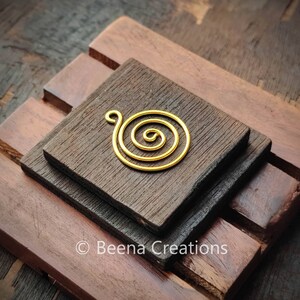 May include: A gold-colored spiral pendant with a loop at the top rests on a dark brown wooden surface. The spiral design is centered and appears to be made of wire. The wooden surface is composed of three stacked, rectangular blocks.