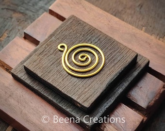 May include: A gold-colored spiral pendant with a loop at the top rests on a dark brown wooden surface. The spiral design is centered and appears to be made of wire. The wooden surface is composed of three stacked, rectangular blocks.