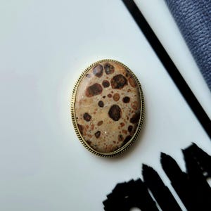 May include: An oval-shaped brooch featuring a polished, tan-coloured stone with dark brown, circular, and irregular spots. The stone is encased in a thin, gold-toned metal frame with a delicate, twisted rope-like texture along the outer edge.