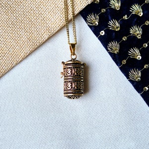 May include: A gold-colored pendant necklace featuring a cylindrical prayer wheel design. The pendant has intricate detailing and is suspended from a delicate chain. The background includes textured fabric and a dark blue velvet material.