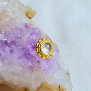 以下が含まれることがあります： クリアな丸い宝石を中央に配した、ゴールドカラーのスタッドピアス。ピアスはスカラップエッジで、紫と白のクリスタルの背景にセットされています。