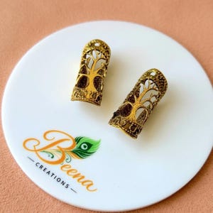 May include: Two cylindrical hair beads in an antique gold finish, each featuring an intricate cut-out design of a tree with spreading branches and roots. The beads are positioned on a white circular surface with the text Beena Creations.