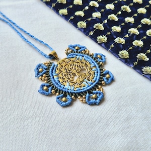May include: A blue and green macrame necklace with a gold tree of life pendant. The pendant is circular and features a detailed design of a tree with branches and leaves.