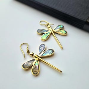 May include: A pair of gold-colored dragonfly earrings. Each earring features a dragonfly design with iridescent wings in shades of green, blue, and gray. The earrings have a hook closure.