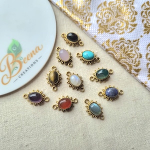 May include: A collection of ten oval-shaped jewellery connectors featuring various cabochon gemstones set in gold-toned metal frames with small loops on each side. The stones include black, pink, turquoise, tiger's eye, white, green, purple, orange, blue, and grey.