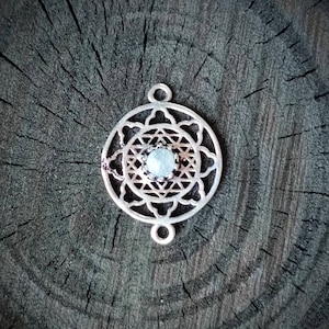 May include: Silver pendant with a geometric design and a clear gemstone in the center. The pendant is on a wooden surface.