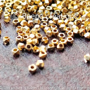 Peut inclure: Gros plan sur un tas de perles en métal de couleur dorée. Les perles sont petites et ont une forme facetée, semblable à un diamant. Elles sont éparpillées sur une surface brune.