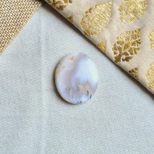 Puede incluir: Un cabujón redondo de ágata blanca con marcas marrones y grises. La piedra tiene una superficie lisa y pulida.