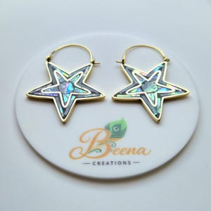 May include: A pair of star-shaped earrings with gold-colored hoops. The stars have a black outline and are filled with iridescent, abalone-like material. The earrings are displayed on a white surface with the text "Beena Creations".