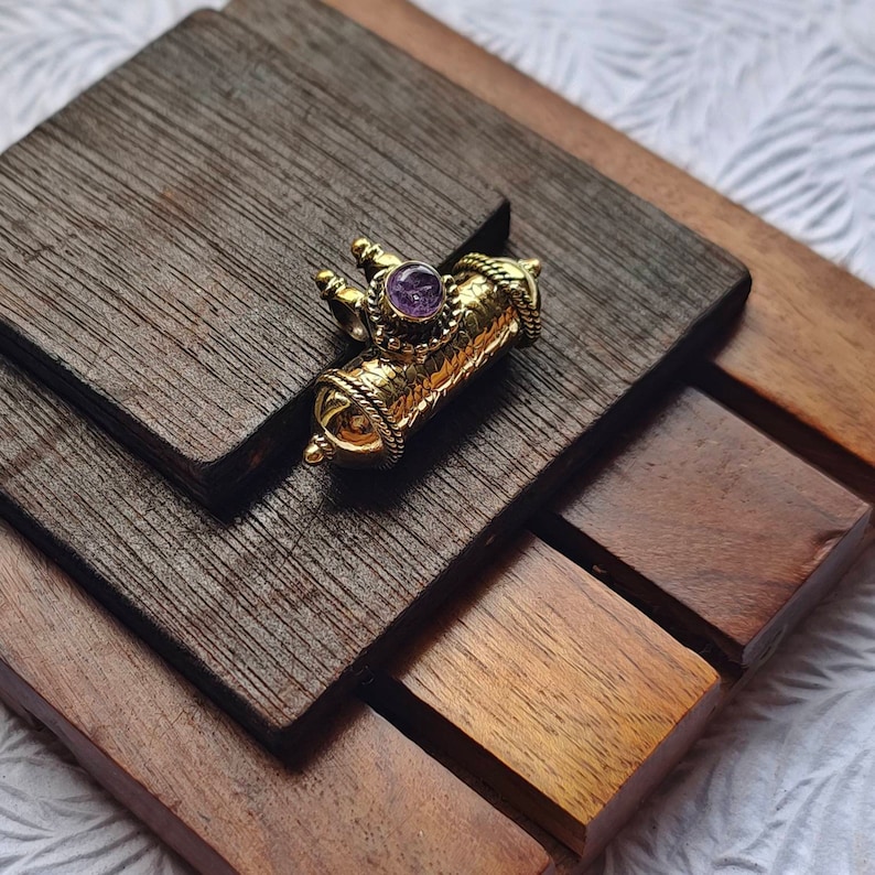 May include: A gold-colored pendant with an intricate design and a purple gemstone in the center. The pendant is shaped like a small cylinder and has a textured surface.
