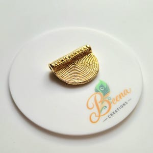 以下が含まれることがあります： 金色の半円形のペンダント。表面はテクスチャ加工が施され、上部は円筒形です。ペンダントは「Beena Creations」の文字が入った白い表面に置かれています。