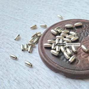 Peut inclure: Gros plan sur un tas de perles en métal argenté sur une surface en bois. Les perles sont de forme ovale et ont une finition lisse et brillante. Les perles sont dispersées autour d'une petite assiette en bois avec le texte "BEENA CREATIONS" dessus.