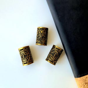 May include: Three cylindrical metal beads with a gold-tone finish and black recessed detailing. Each bead features an intricate embossed pattern of interconnected lines and circular motifs, creating a textured surface across the entire exterior.