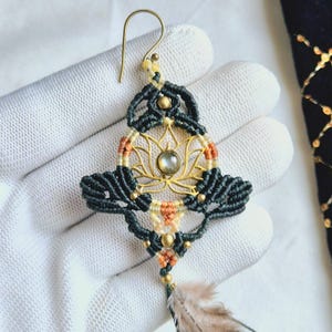 May include: Green and gold macrame earrings with a lotus flower design and a feather charm. The earrings feature a light blue gemstone in the center of the lotus flower.