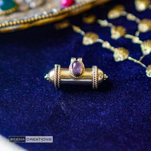 Puede incluir: Un colgante de plata y oro con una piedra preciosa púrpura en el centro. El colgante tiene forma de un pequeño cilindro y presenta detalles intrincados.