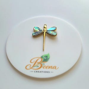 Könnte beinhalten: Ein goldfarbener Libellenanhänger mit schillernden Abalone-Muscheleinlagen in den vier Flügeln. Der Anhänger befindet sich über dem Schriftzug Beena CREATIONS, der eine stilisierte Pfauenfedergrafik in Grün- und Orangetönen enthält.