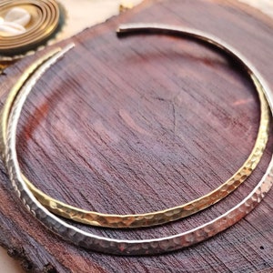 May include: Two hammered metal bangles, one silver and one gold, are stacked on a wooden surface. The bangles are both round and have a textured finish.