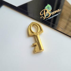 May include: A small, gold-colored key with an arched top and a circular opening. The key has a detailed design and is set against a white surface with a black card that has the text "Beena Creations".