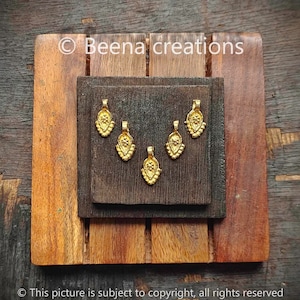 May include: Five gold-colored metal charms in the shape of a teardrop with a floral design. The charms are arranged in a semi-circle on a dark brown wooden surface.  © Beena creations