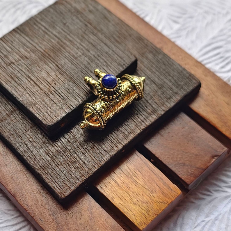 May include: A gold-toned pendant with intricate details and a blue gemstone in the center. The pendant is on a wooden surface.