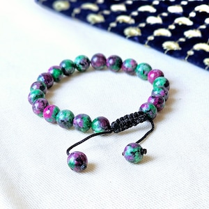 May include: A green and pink gemstone bracelet with a black cord and adjustable clasp. The bracelet is made of round beads.