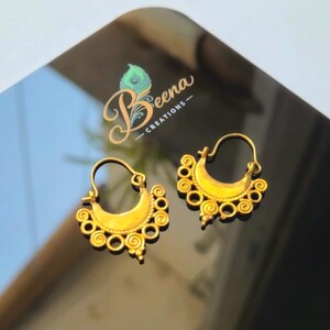 May include: A pair of gold-colored hoop earrings with intricate spiral and circular designs. The earrings have a crescent shape and are displayed on a reflective surface. The brand name "Beena Creations" is visible in the background.