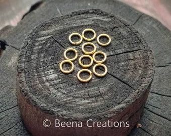 Puede incluir: Primer plano de un grupo de 15 anillos de salto dorados sobre una superficie de madera oscura. Los anillos de salto son pequeños y redondos, y están dispuestos en una pila suelta.