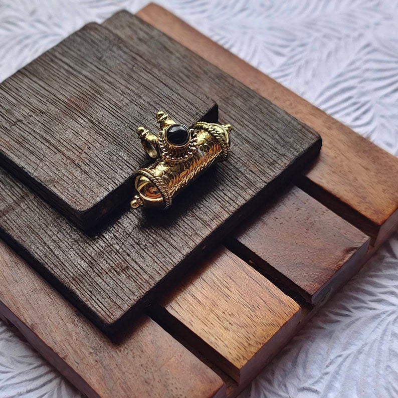 May include: A gold-toned pendant with a black stone in the center. The pendant is in the shape of a cylinder with intricate details and a decorative top.