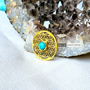 May include: A gold-colored metal pendant featuring a flower of life design with a turquoise gemstone in the center. The pendant is resting on a piece of clear quartz.