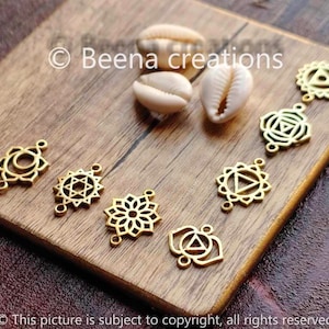 Może przedstawiać: Siedem złotych metalowych zawieszek o różnych wzorach, w tym kwiat, gwiazda i trójkąt. Zawieszki leżą na drewnianej powierzchni z dwoma muszelkami.