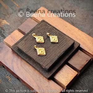 May include: Three gold-coloured square pendants with intricate designs. The pendants are on a wooden surface.